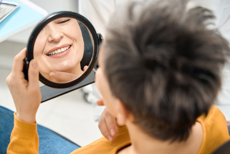 A dental patient smiles while looking at their reflection in a handheld mirror, admiring their newly restored teeth during a dental visit.