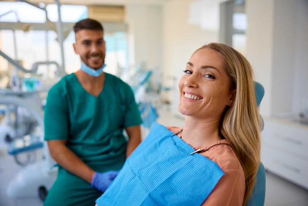 A woman sits in a dental chair smiling at the camera, wearing a blue dental bib. Behind her, a dentist in green scrubs with a face mask around his neck is seated, smiling. Dental equipment is visible in the bright room.